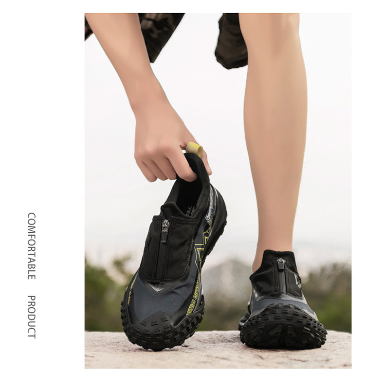 A person holding a pair of black outdoor casual shoes with a zipper, positioned on a rock in an outdoor setting with greenery, emphasizing the shoes' comfortable and practical design for outdoor use.