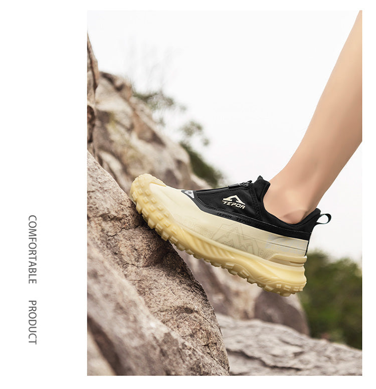 A person wearing black and yellow outdoor sports shoes, stepping on a rock in a natural outdoor setting with greenery, highlighting the shoes' comfortable and grippy design for outdoor activities.