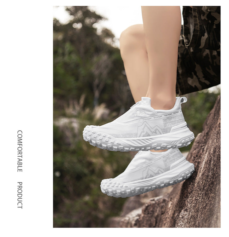 A person wearing white outdoor casual shoes, positioned on a rock in a natural outdoor setting with greenery, highlighting the shoes' comfortable and practical design for outdoor activities.